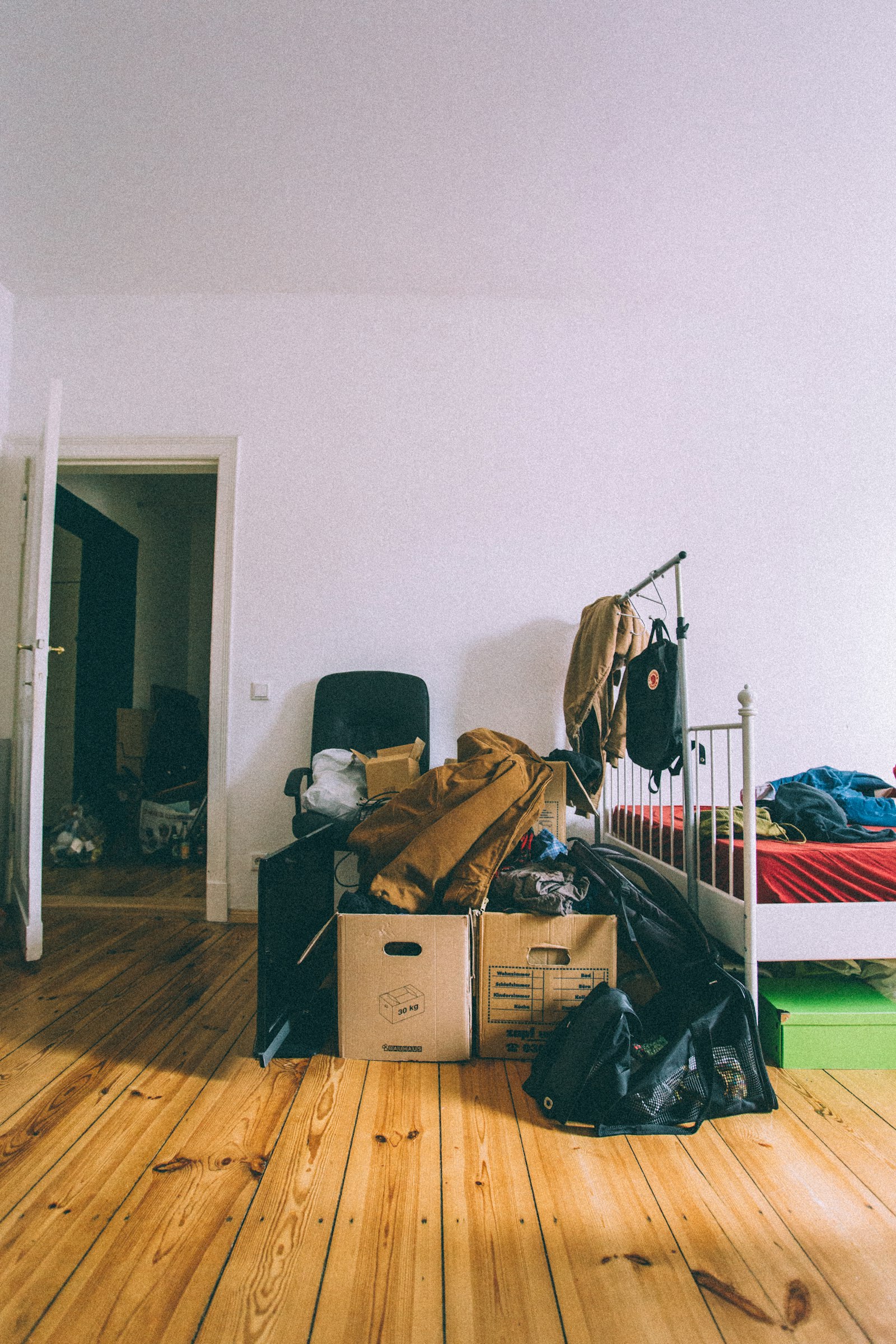 A half-packed bedroom with two open moving boxes on a bare wooden floor, coats piled on top and a bed pushed against the wall.