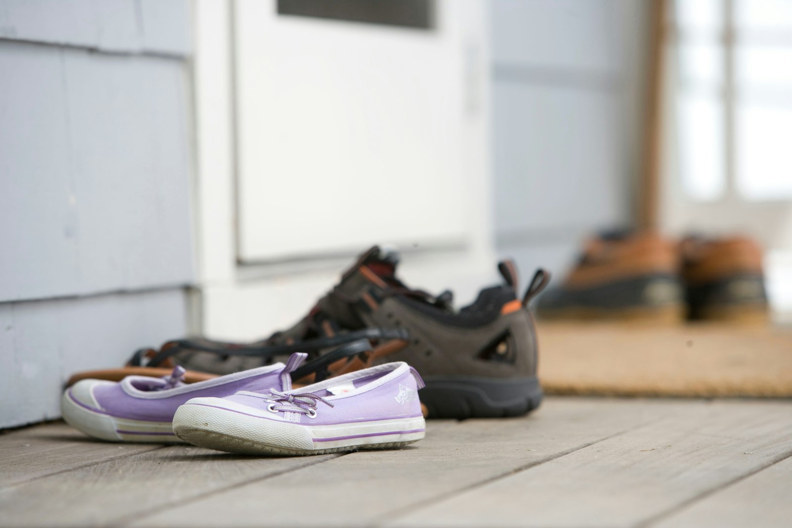 A child's purple canvas shoes on a porch board beside two pairs of adult work shoes, next to a screen door.