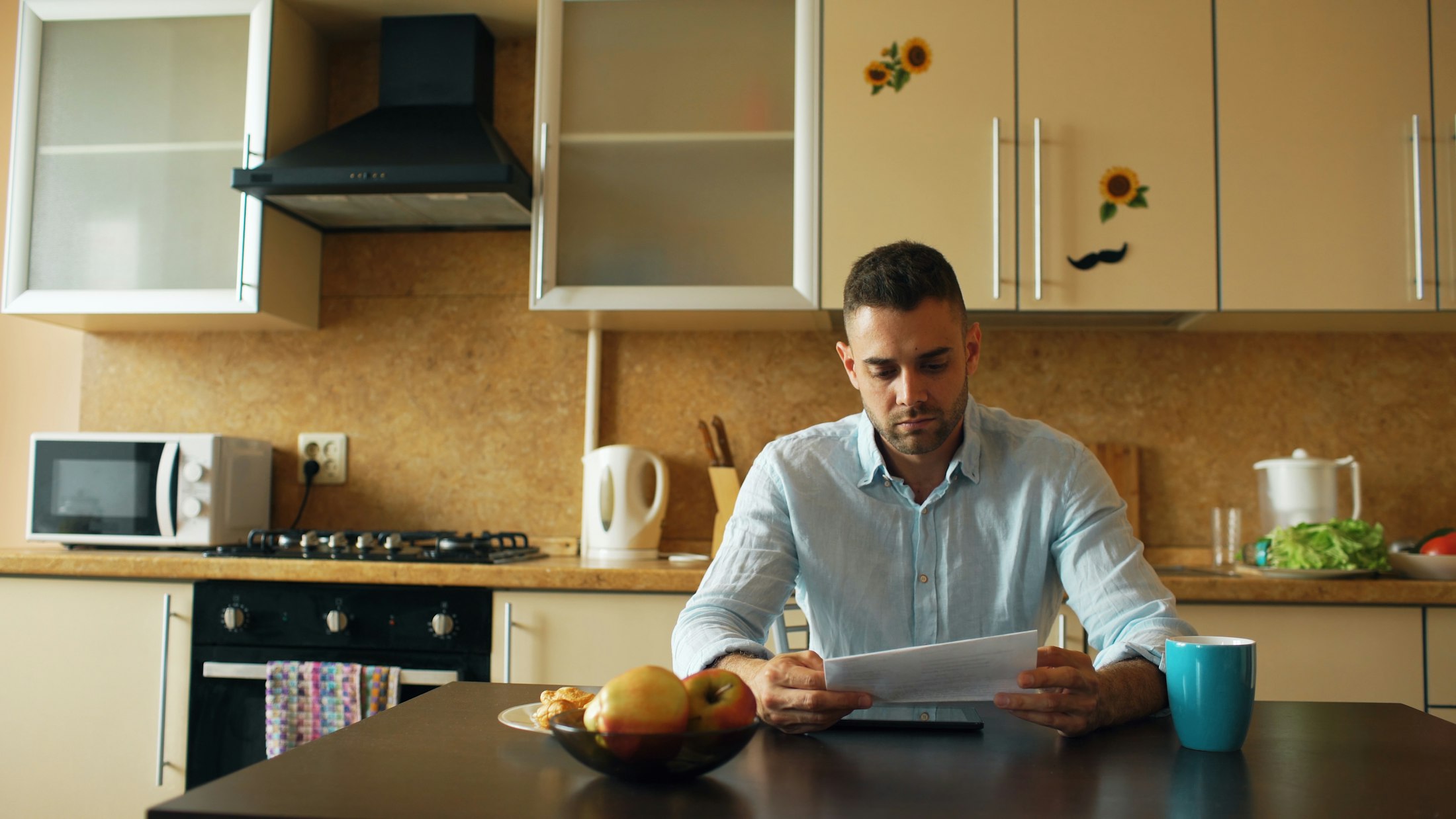A man sitting at a kitchen table in the morning, holding a letter with both hands and reading it.